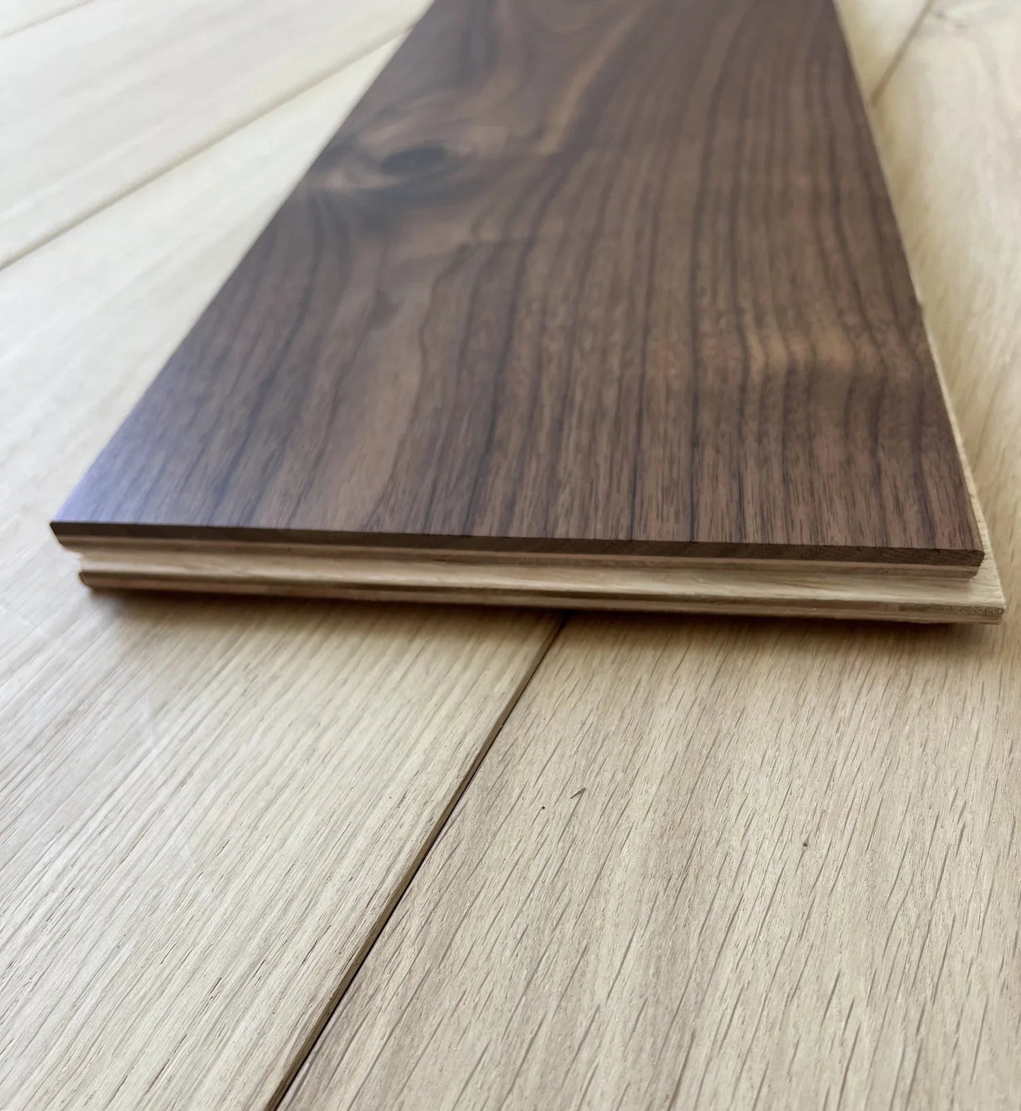 A close-up of a dark walnut wood flooring plank resting on a light oak wood floor, showing the wood grain and layered construction.