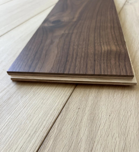 A close-up of a dark walnut wood flooring plank resting on a light oak wood floor, showing the wood grain and layered construction.