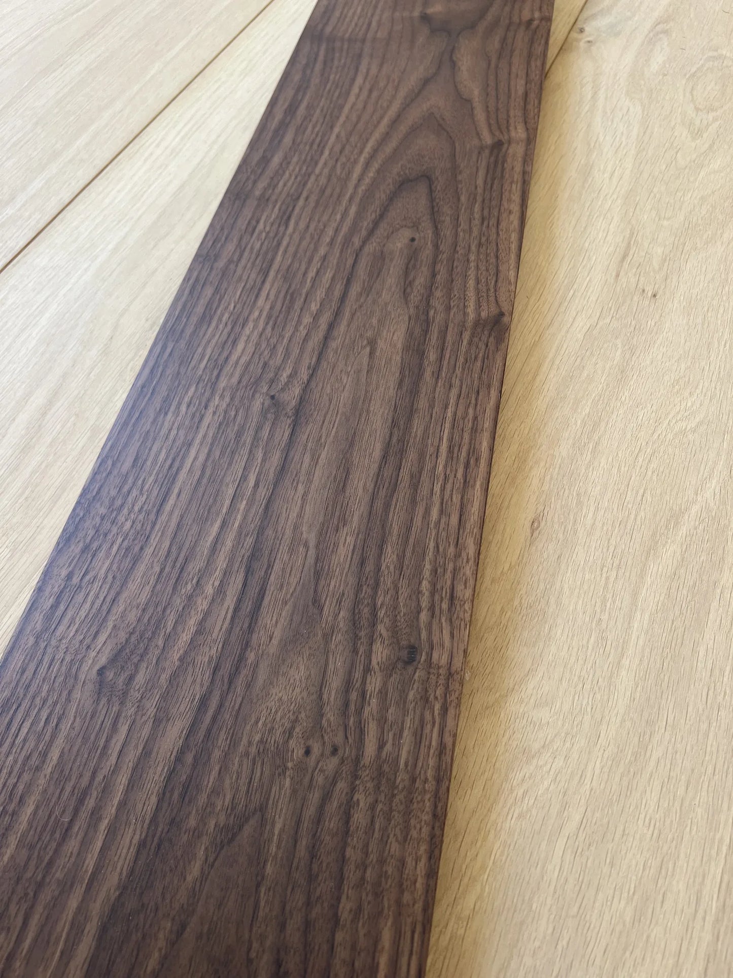 Walnut wood plank with prominent grain patterns, shown diagonally on a light oak wood floor background.